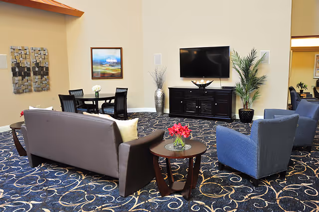 A senior living facility common area with a brown leather sofa, two blue armchairs, a round wooden side table with a vase of red flowers, a dark wood TV stand with a flat-screen TV mounted above it, decorative plants, a round dining table with four chairs, and wall art. The room has a patterned blue carpet and beige walls.
