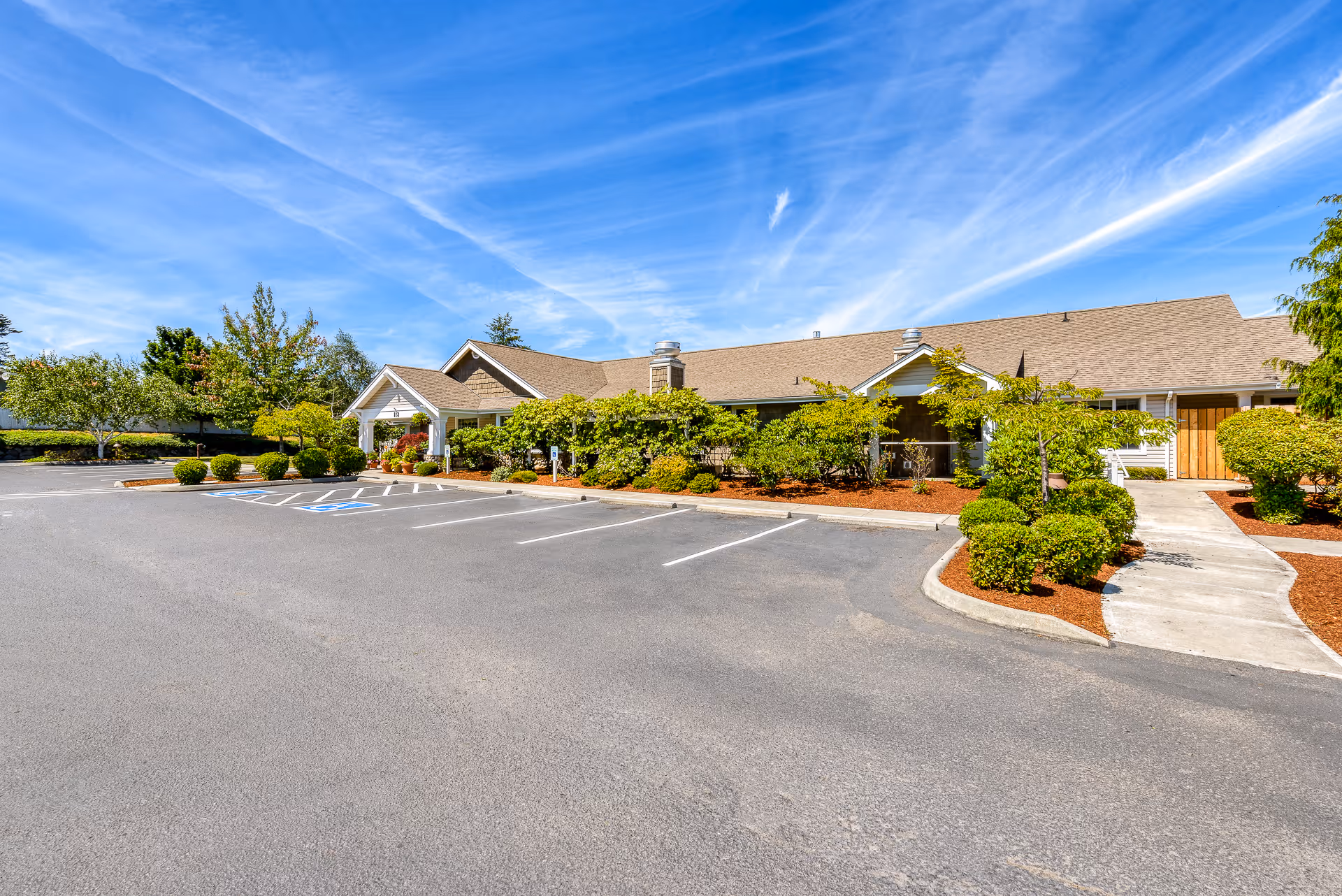 Single-story memory care facility building front with landscaped entrance and an empty parking lot under a bright blue sky.