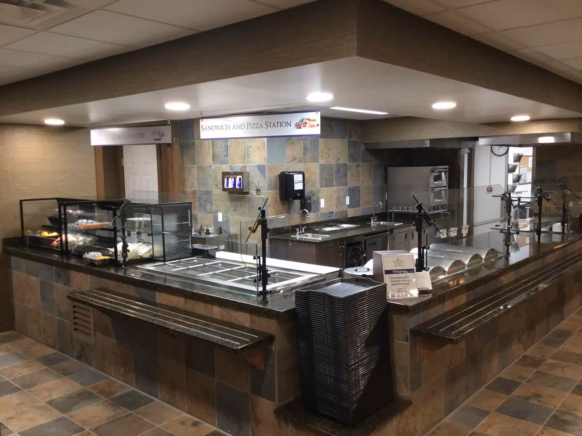A food service counter area in a retirement community with a sandwich and pizza station sign overhead. The counter has trays, a stack of black trays for carrying food, and a glass display case with food items. The background shows a kitchen area with stainless steel appliances and tiled walls.