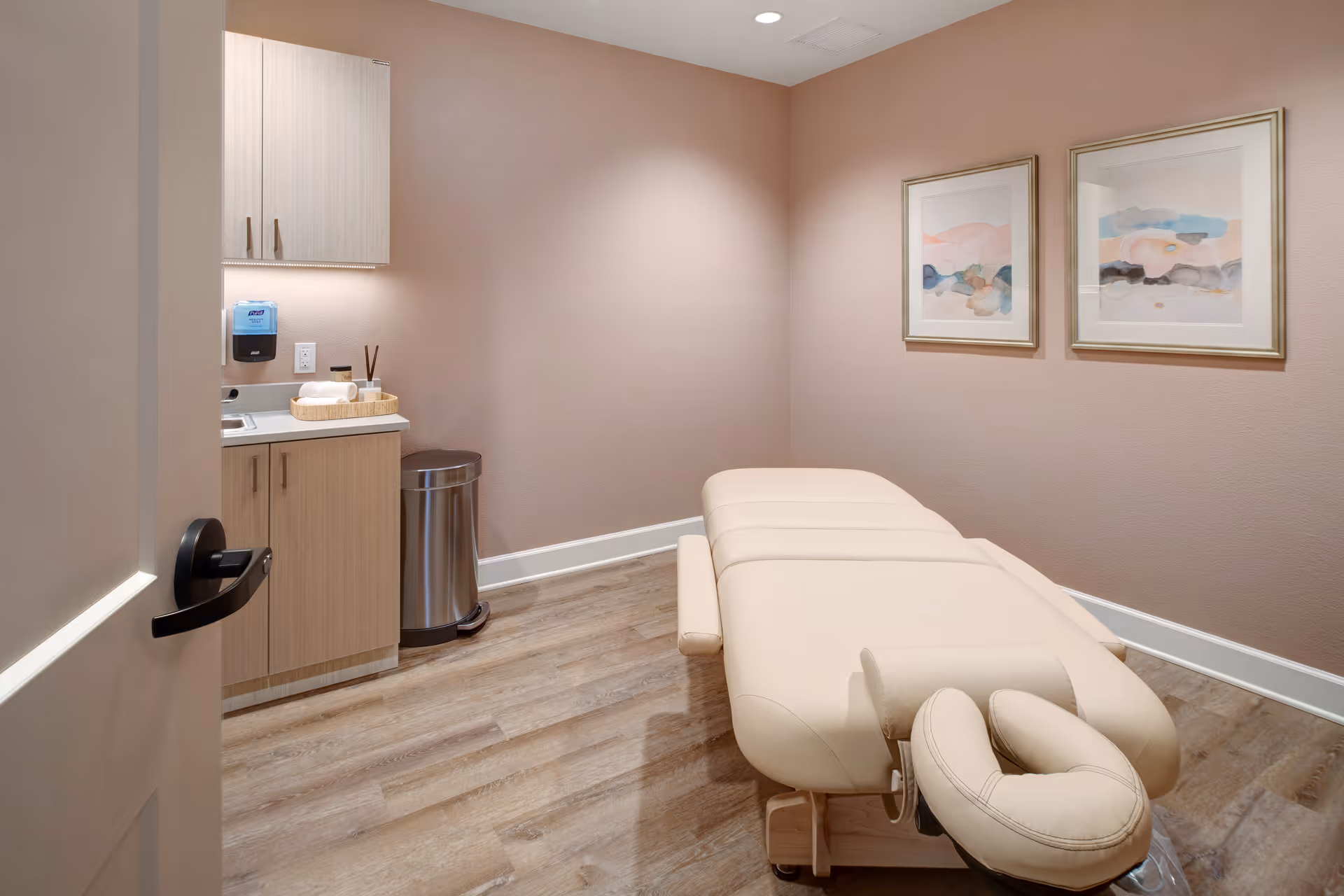 Massage therapy room with a beige treatment table, sink and cabinets, and two framed prints on pink walls.