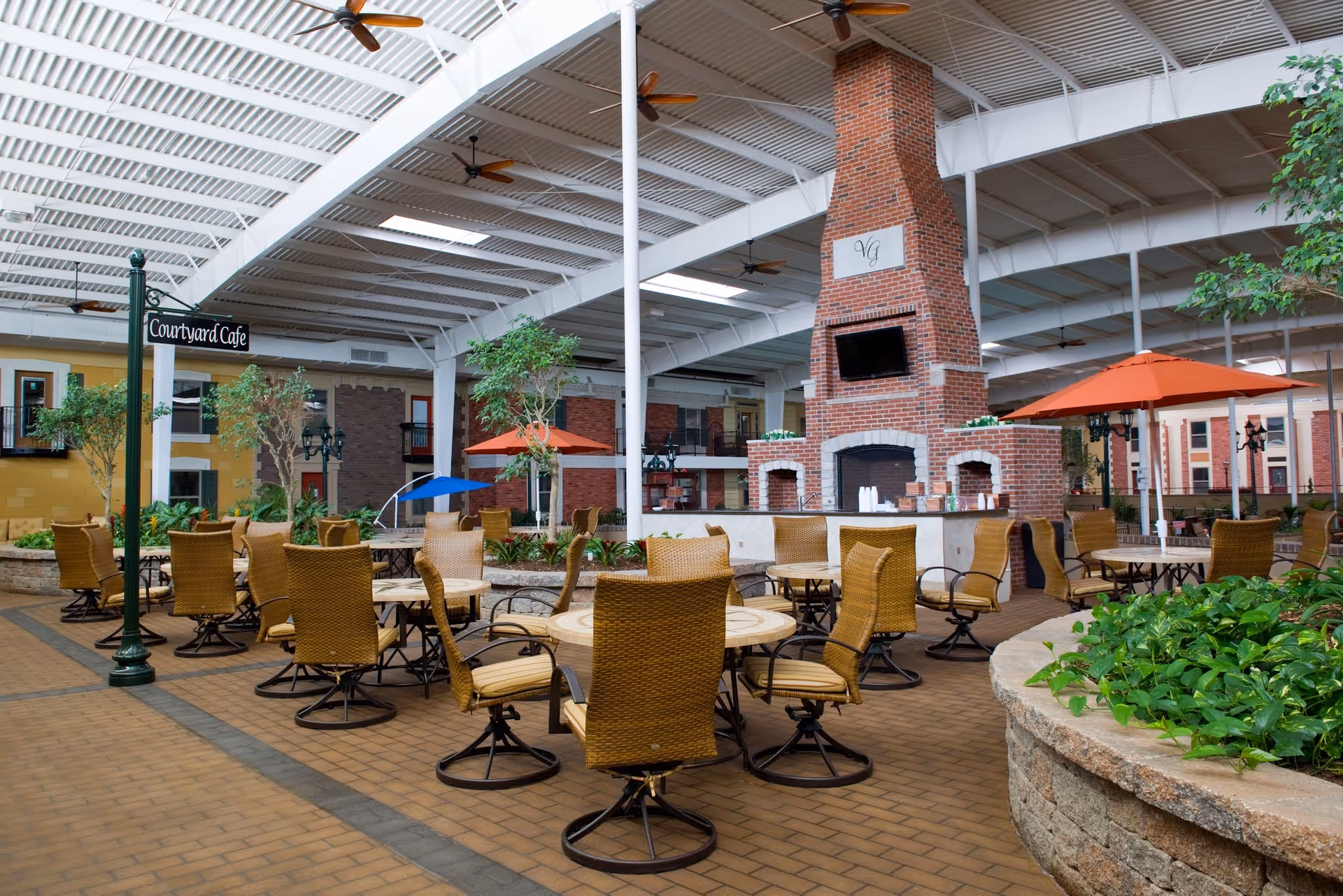 Indoor courtyard cafe area with multiple round tables and wicker chairs arranged around them. There are green plants in stone planters, several orange umbrellas, and a large brick fireplace with a TV mounted above it. The ceiling is white with exposed beams and ceiling fans. A sign reads 'Courtyard Cafe'.