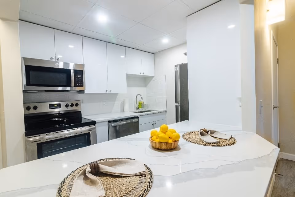 Modern kitchen with white cabinets, stainless steel appliances including a microwave, stove, dishwasher, and refrigerator. A white marble countertop island with two woven placemats and napkins, and a bowl of lemons is in the foreground.