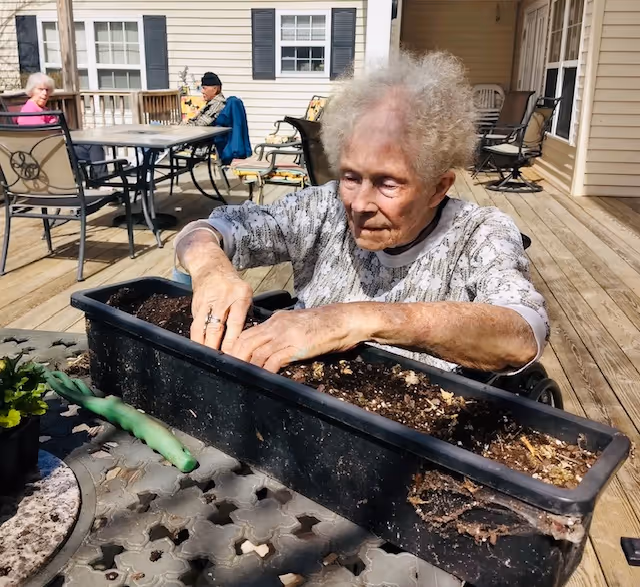 An elderly woman with curly white hair is sitting outdoors on a wooden deck, planting or tending soil in a long black planter box placed on a table. In the background, two other elderly individuals are seated at a table near a beige building with windows and black shutters. The scene is sunny and appears to be a communal outdoor area of a care facility.