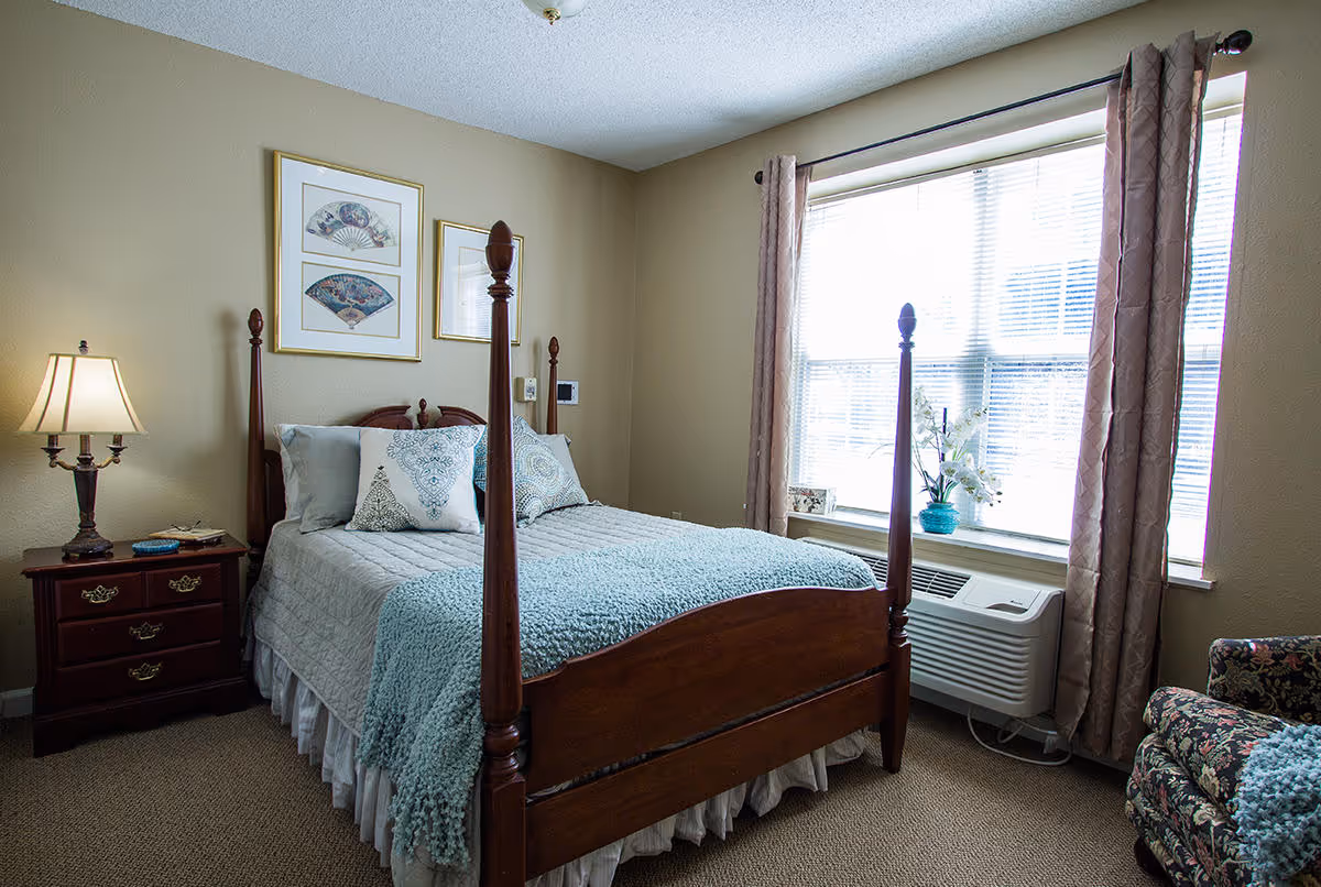 A cozy bedroom with a wooden four-poster bed made with light blue and white bedding and decorative pillows. There is a wooden nightstand with a lamp on the left side of the bed. Two framed pictures hang on the beige wall above the bed. A large window with blinds and light brown curtains lets in natural light. Below the window is an air conditioning unit with a blue vase and white flowers on the windowsill. A floral patterned armchair is partially visible on the right side of the image.