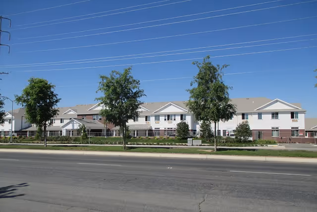 Exterior view of a large two-story senior living facility building with white and brick facade, surrounded by a few trees and green landscaping under a clear blue sky.