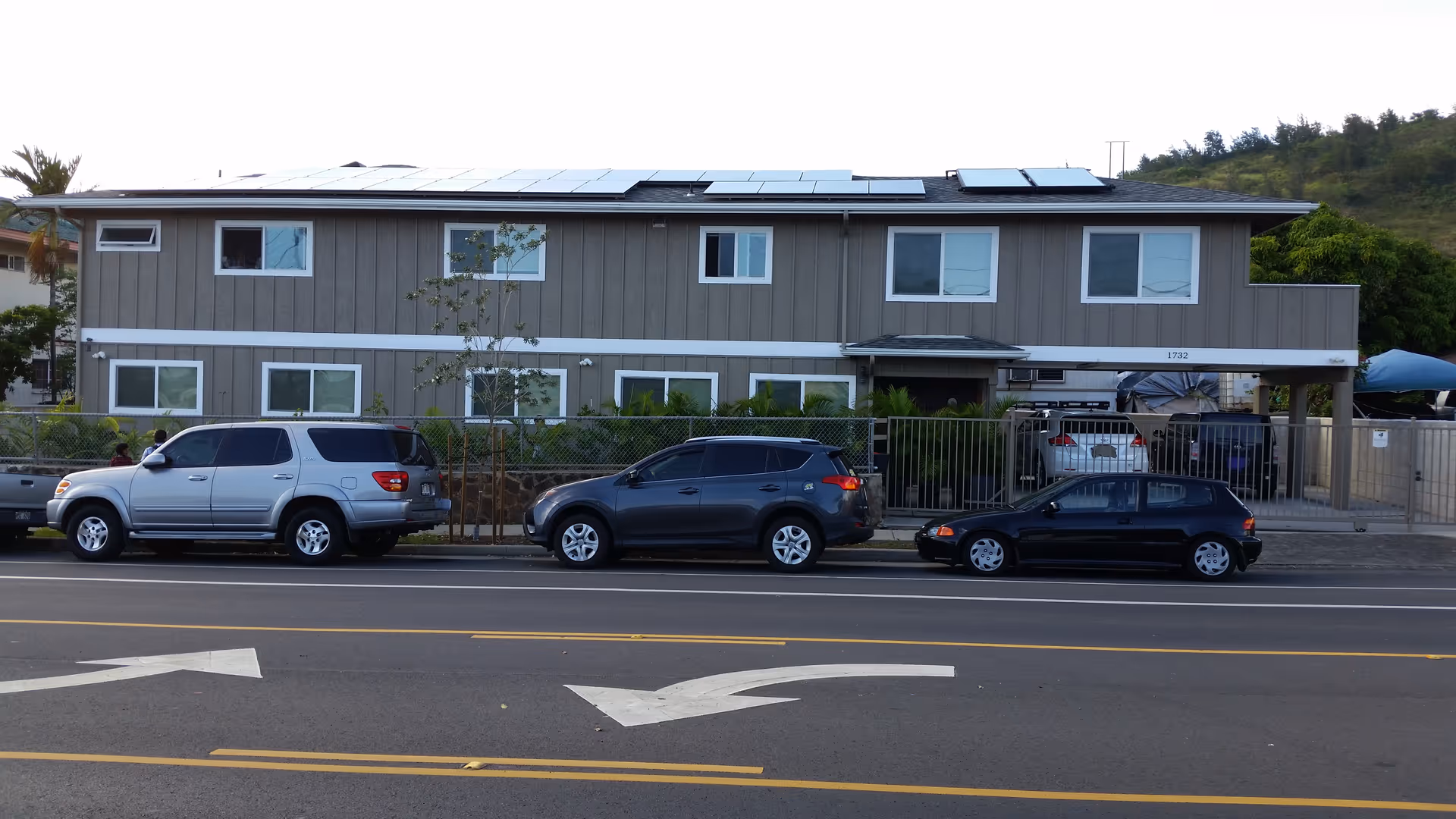 Exterior view of a two-story gray building with multiple windows and solar panels on the roof. There are three cars parked along the street in front of the building, which is surrounded by a metal fence. The building is located on a paved road with lane markings and directional arrows.