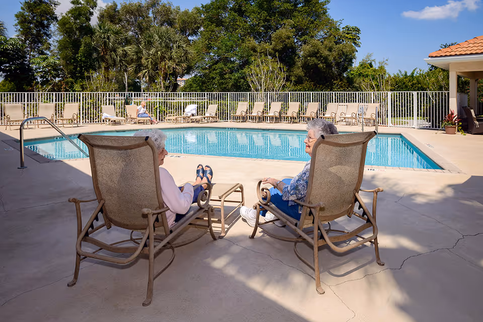 Two older women seated on lounge chairs talking beside an outdoor swimming pool at a senior living facility.
