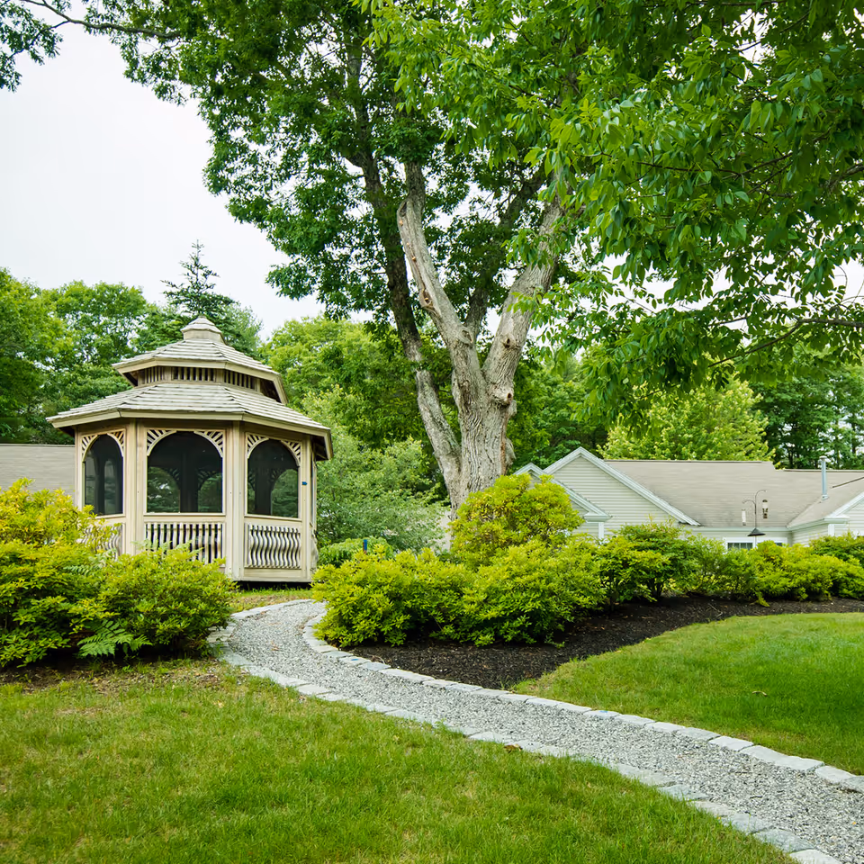 A peaceful outdoor garden area with a gravel pathway leading to a wooden gazebo surrounded by lush green bushes and trees, with a building partially visible in the background.