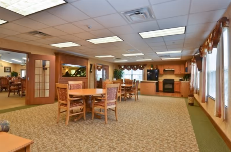 Carpeted communal dining area with round tables and chairs and a kitchen area in the background.