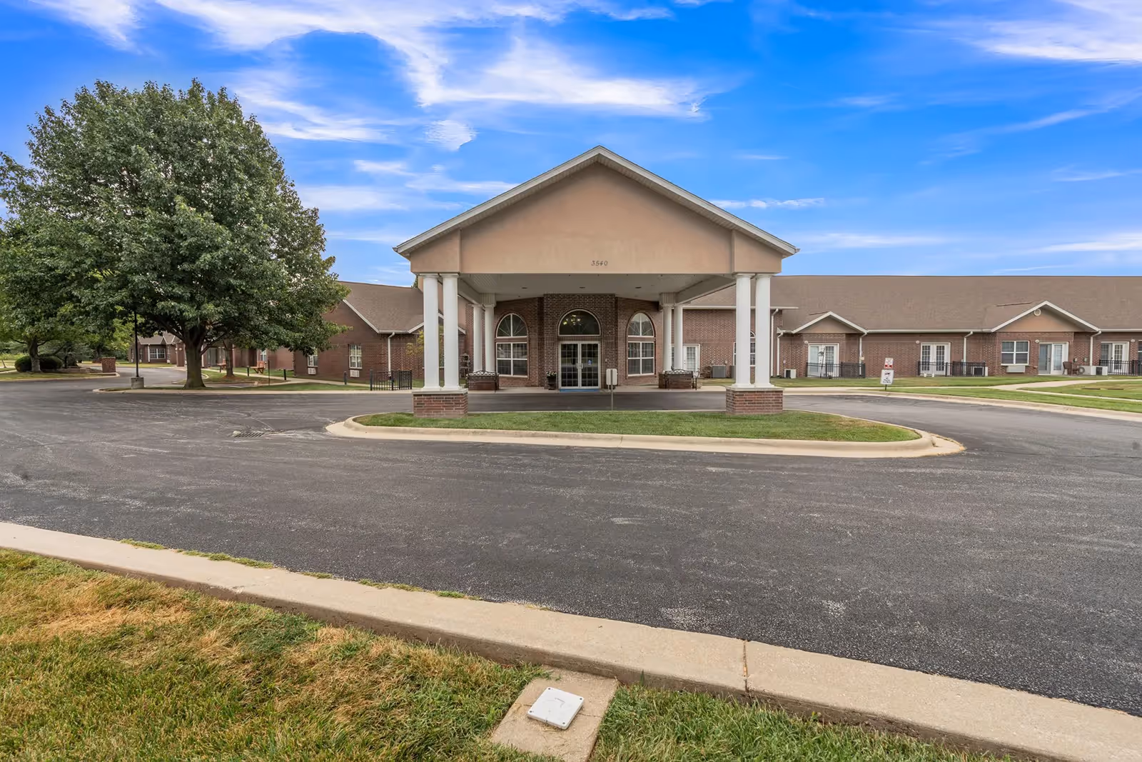 Front exterior view of a single-story senior living facility building with a covered entrance supported by white columns, surrounded by a paved driveway and green lawns under a blue sky with scattered clouds.