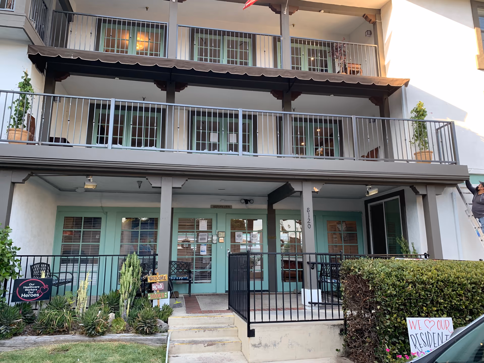 Front exterior view of a two-story assisted living facility with green doors and windows, a balcony with railings on the second floor, potted plants, and signs expressing appreciation for healthcare workers and residents.