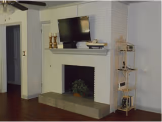 Living room with a white painted brick fireplace and mantel holding a TV and decorative items, hardwood floors, and a tall shelving unit to the right.