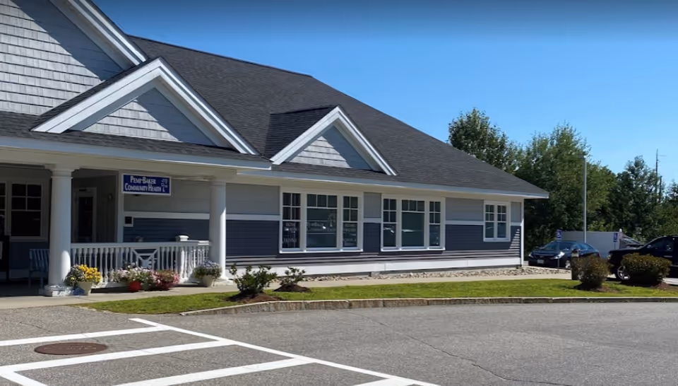 Exterior view of a single-story building with gray siding and white trim, featuring a covered porch with white columns and a sign that reads 'Pemi-Baker Community Health'. There are several windows along the side of the building, a small landscaped area with bushes and flowers, and a parking lot with marked spaces in front.