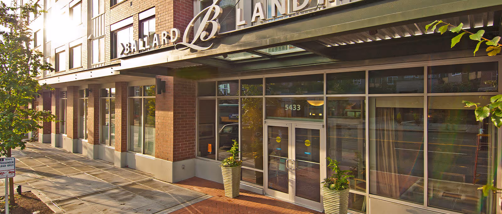 Exterior view of the entrance to Ballard Landmark building with large glass doors, brick walls, and a metal awning above the entrance. Two large planters with green plants flank the doorway. The sidewalk and street are visible with some trees and a sign indicating a 3-minute passenger load zone.