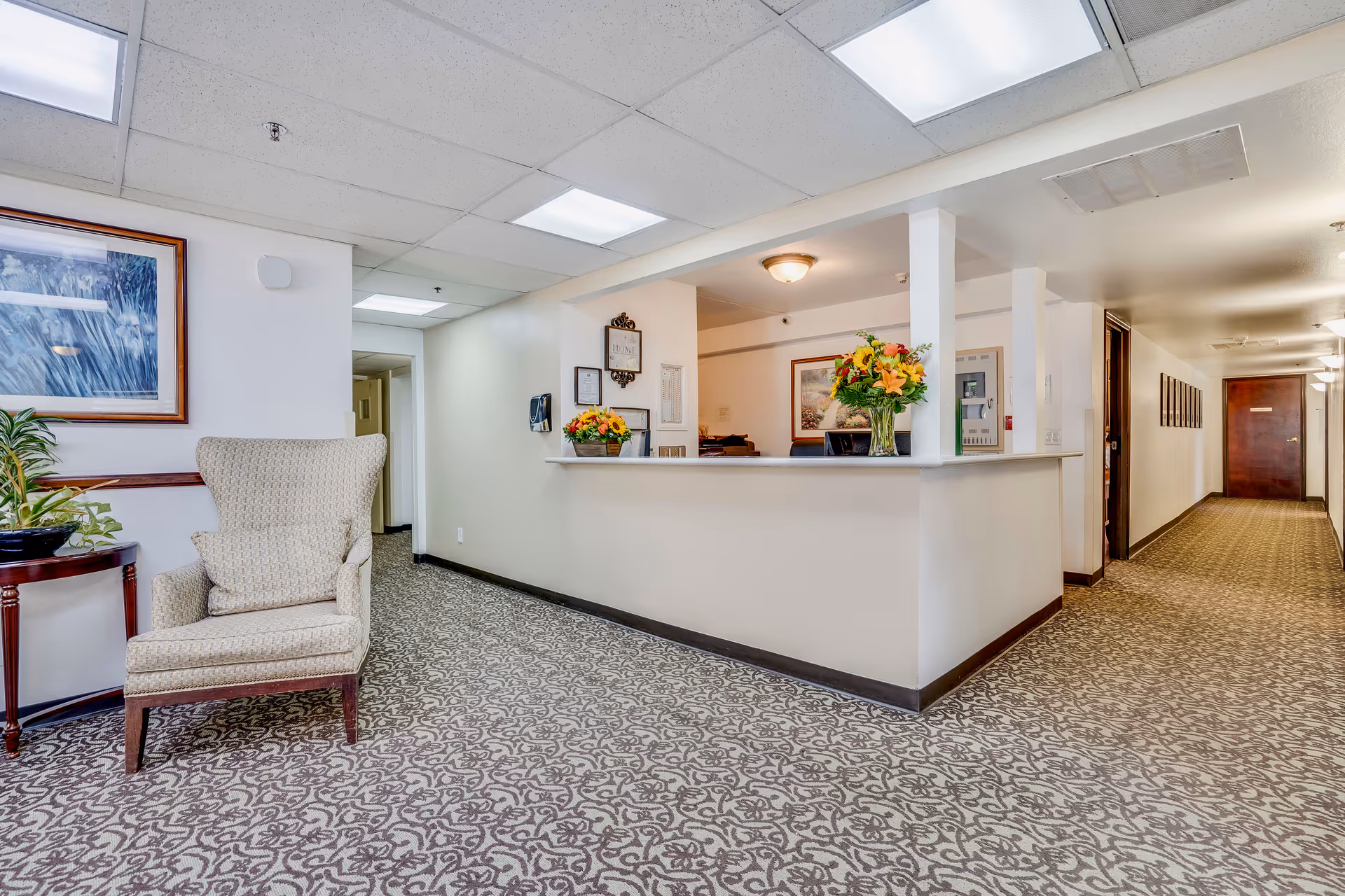 A carpeted assisted living reception area with a front desk, floral arrangements on the counter, and a cushioned armchair by a hallway.