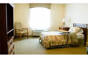 A bright senior living bedroom with a single bed, overbed table, nightstand and chair beside a large window.