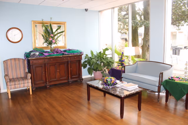 A bright living room area with wooden flooring, a striped armchair on the left, a dark wooden cabinet with a floral arrangement and decorations on top, a large mirror above the cabinet, and a round wall clock. On the right side, there is a light blue upholstered sofa, a marble-top coffee table with magazines and decorative items, a potted plant, and large windows letting in natural light.