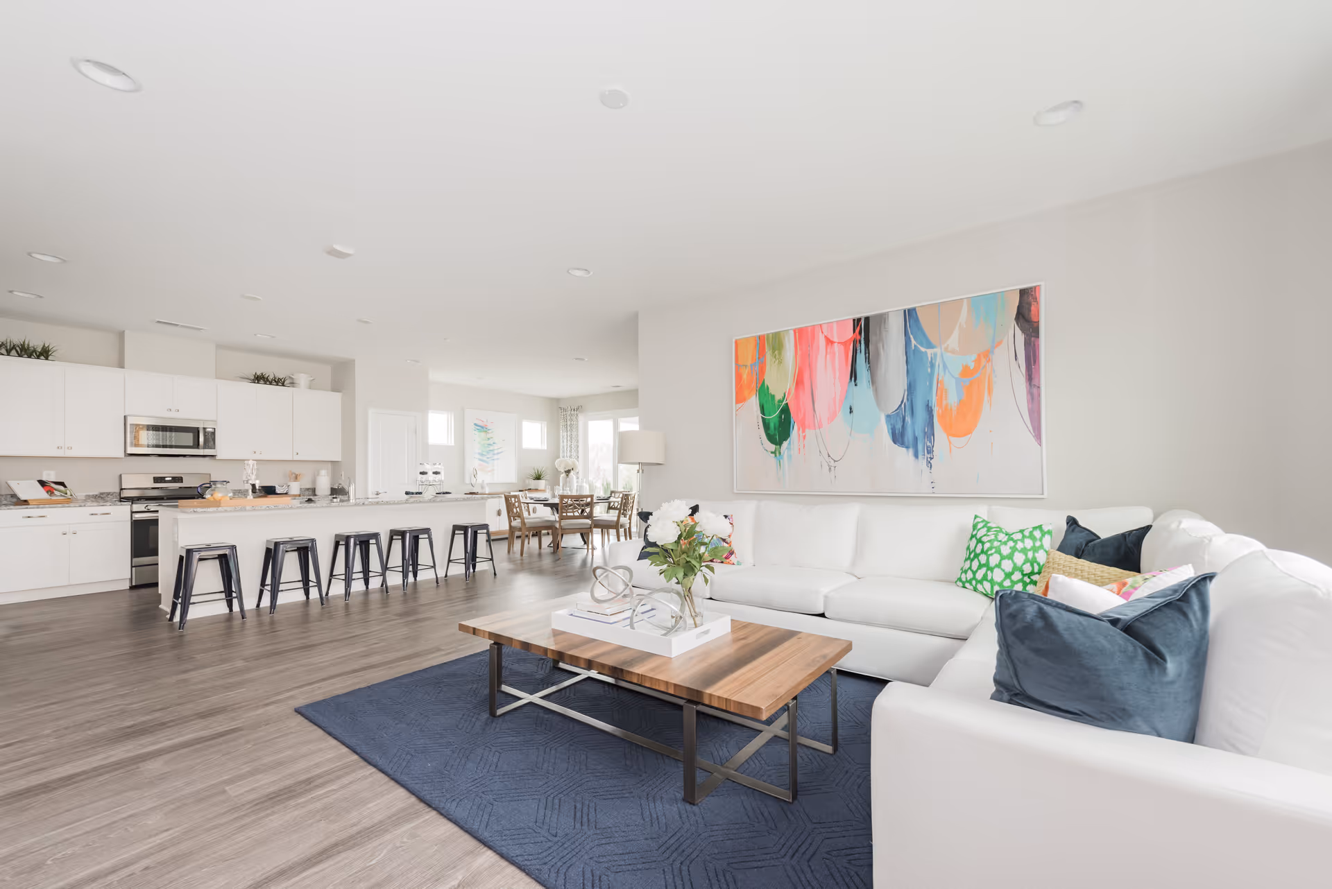 Bright and spacious open-concept living area featuring a white sectional sofa with colorful pillows, a wooden coffee table on a blue rug, and a large abstract painting on the wall. The living area flows into a modern kitchen with white cabinetry, stainless steel appliances, and a long island with black bar stools. In the background, a dining area with a wooden table and chairs is visible near large windows.