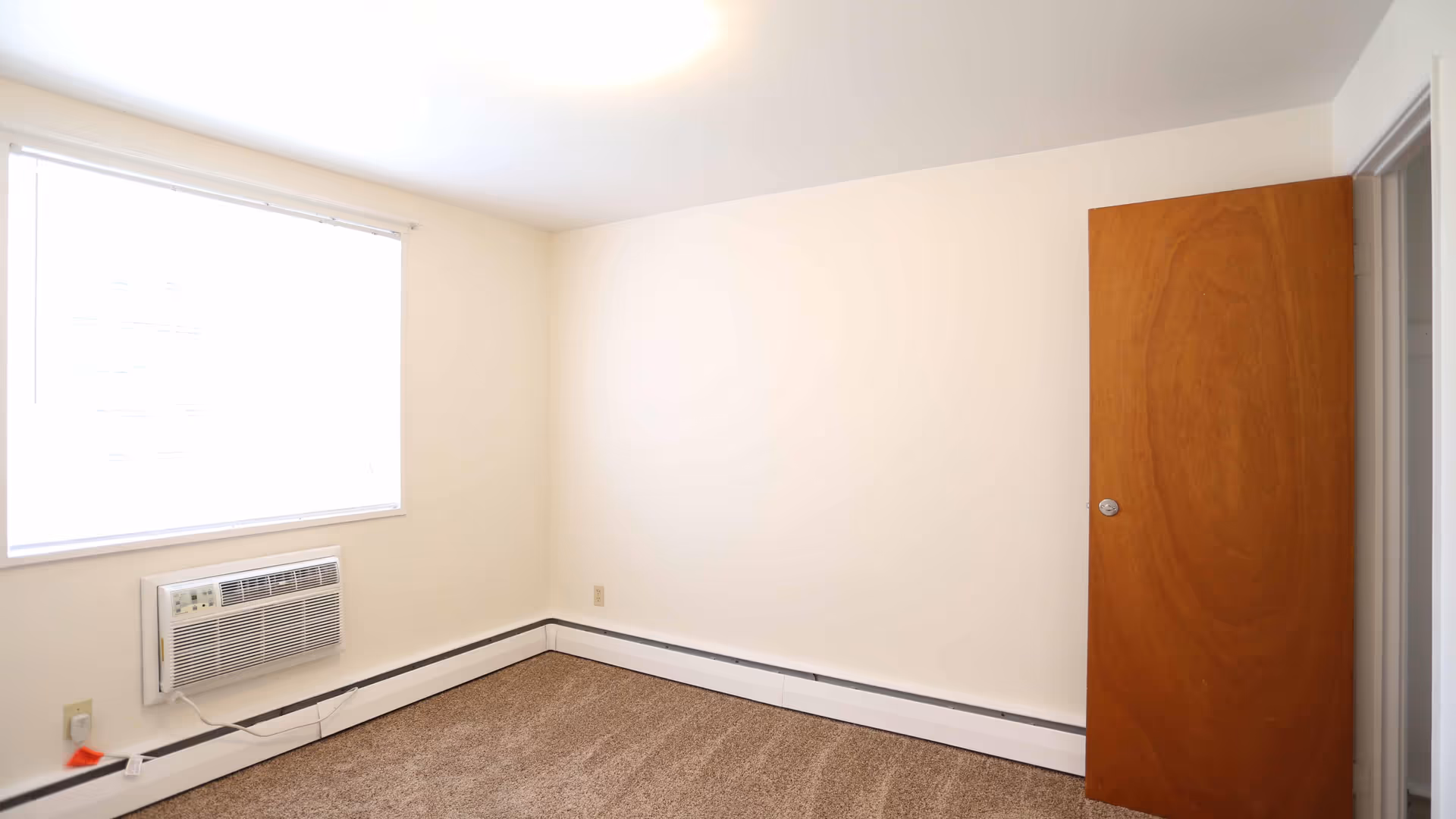 Empty carpeted bedroom with a window air conditioner, cream-colored walls, and a wooden door.