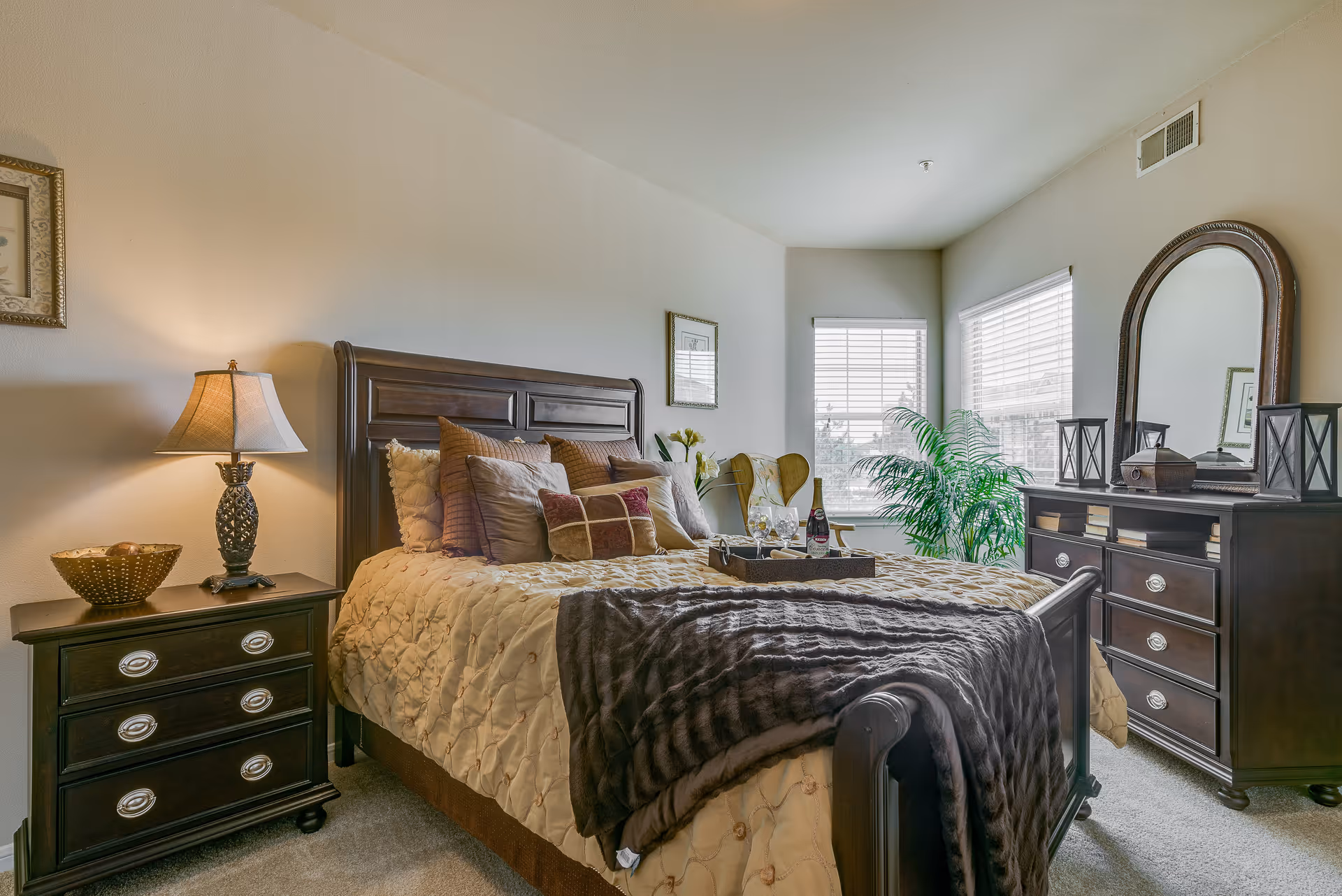 A well-lit bedroom featuring a large wooden bed with multiple pillows and a beige quilt. A dark brown throw blanket is draped across the foot of the bed. To the left is a nightstand with a decorative lamp and a bowl. On the right side of the room is a dresser with a large mirror, two lantern-style candle holders, and a small decorative box. Two windows with white blinds allow natural light to fill the room, and a green potted plant is placed near the windows. The walls are painted a light beige color.