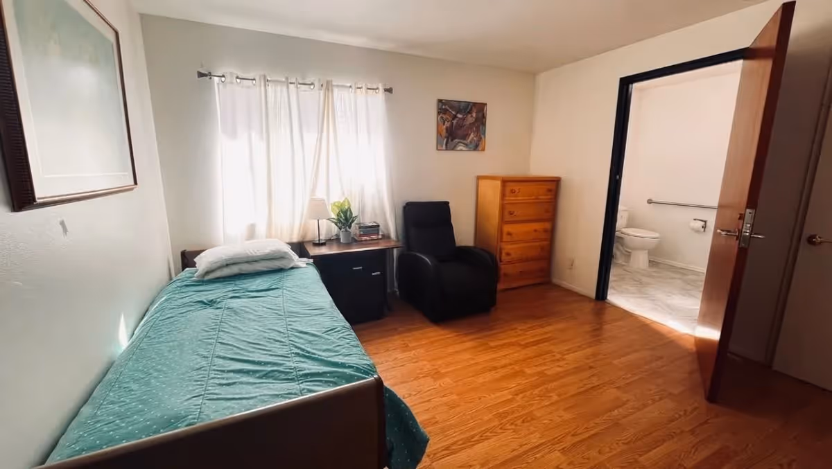 A simple bedroom with a single bed covered in a green bedspread, a nightstand with a lamp and plant, a black armchair, a wooden dresser, and an open door leading to a bathroom with a visible toilet and grab bar.