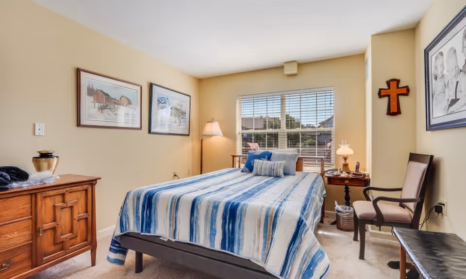 A bedroom with a bed covered in a blue and white striped blanket, two framed pictures on the wall, a wooden dresser, a floor lamp, a side table with a lamp and items on it, a chair, a wooden cross on the wall, and a large window with blinds letting in natural light.