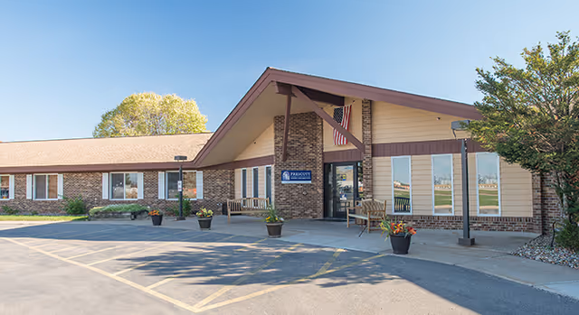 Exterior view of Prescott Nursing & Rehab building with a brick and beige facade, a peaked roof entrance, several windows, potted plants near the entrance, and an American flag hanging above the door under a clear blue sky.