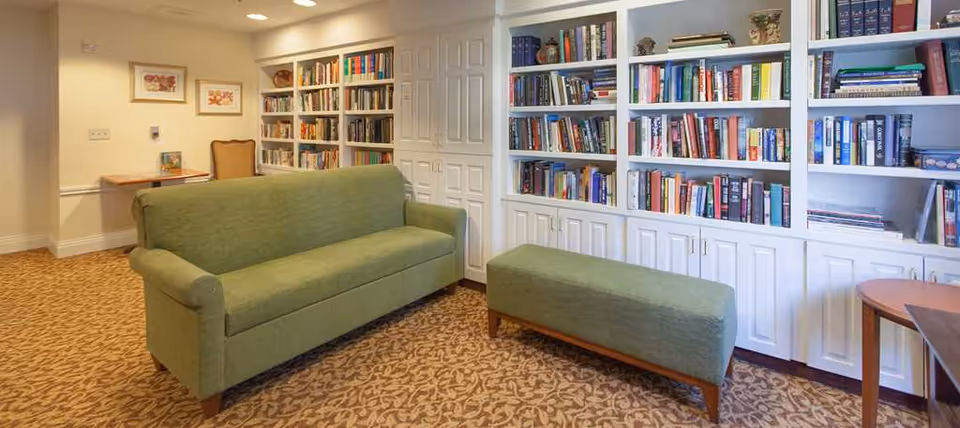 A cozy reading room with a green sofa and matching ottoman placed on a patterned carpet. The room features built-in white bookshelves filled with books and decorative items. There is a small wooden table with a chair in the corner and framed artwork on the wall.