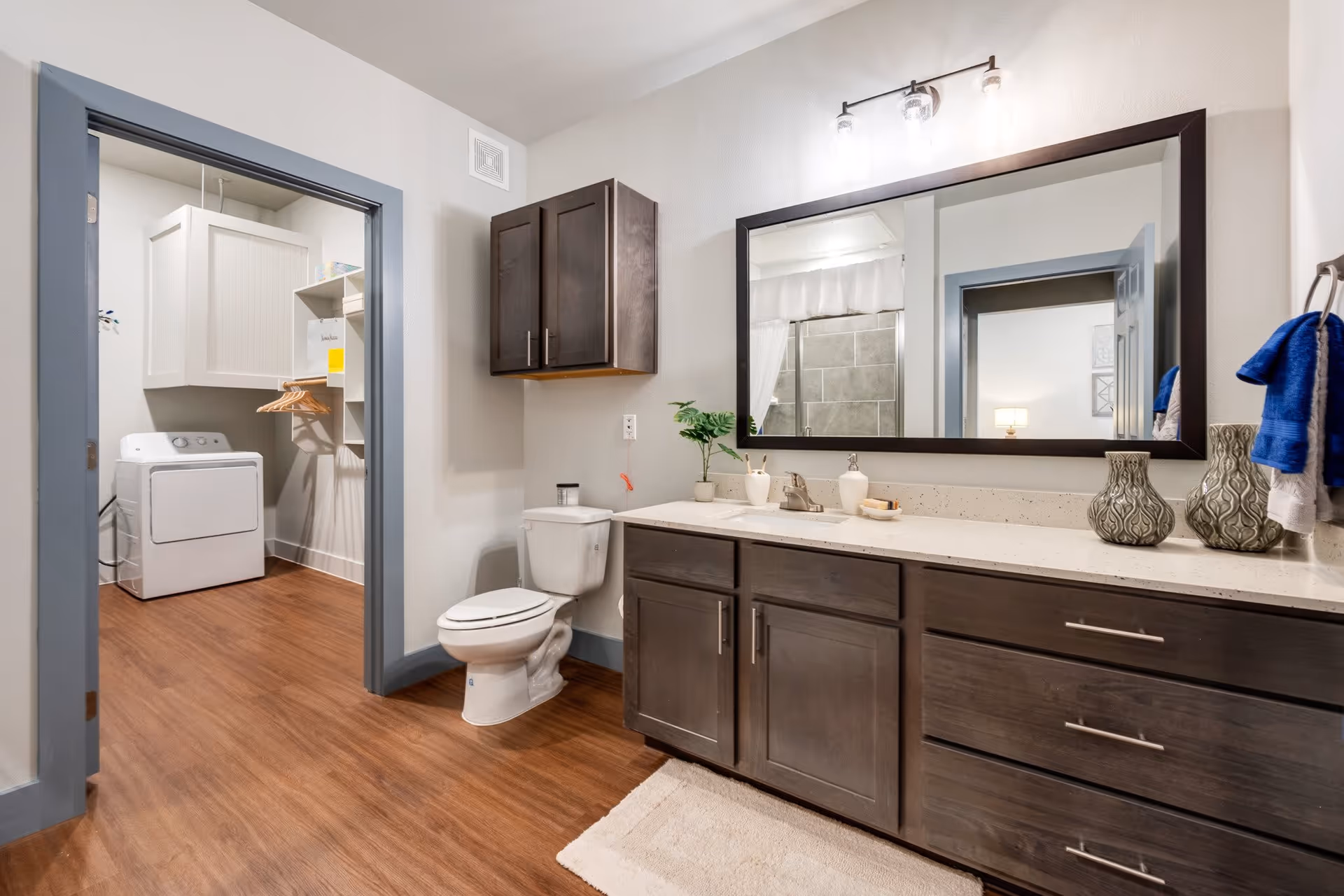 Modern bathroom with a dark wood vanity and large mirror, a toilet, and a visible laundry area with a washer and shelving.