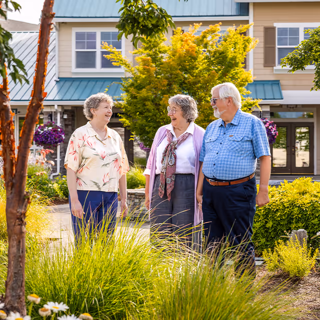 Three elderly people standing outside in a garden area with green plants and flowers, smiling and talking. Behind them is a residential building with beige siding, blue metal roofing, and windows.