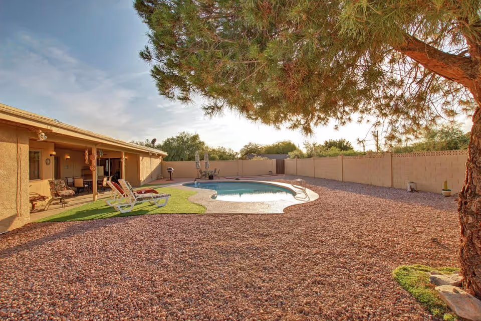 Outdoor area of Sweetwater Groves By Majestic Residences featuring a swimming pool surrounded by a gravel yard and a covered patio with lounge chairs and a dining table. A large tree provides shade in the foreground.