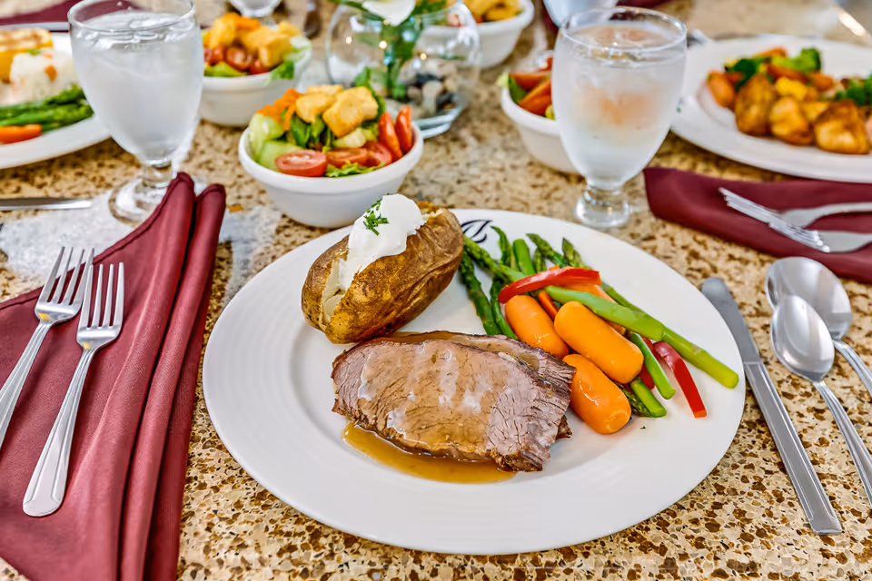 A plated meal consisting of a slice of roast beef with gravy, a baked potato topped with sour cream and chives, and steamed vegetables including carrots, asparagus, and red bell peppers. The plate is set on a speckled countertop with silverware and a maroon cloth napkin beside it. In the background, there are bowls of salad and glasses of water.