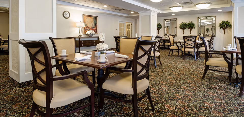 Dining room with multiple wooden tables and upholstered chairs set for meals in an assisted living facility.