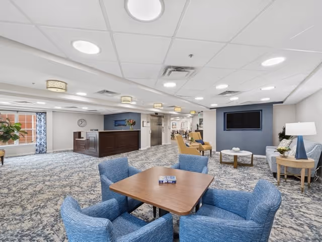 A spacious senior living facility common area with patterned carpet, a wooden table surrounded by four blue upholstered chairs, a seating area with a sofa, armchairs, a coffee table, and a side table with a lamp. There is a large flat-screen TV mounted on a blue accent wall, a reception desk in the background, and large windows with blue patterned curtains letting in natural light.