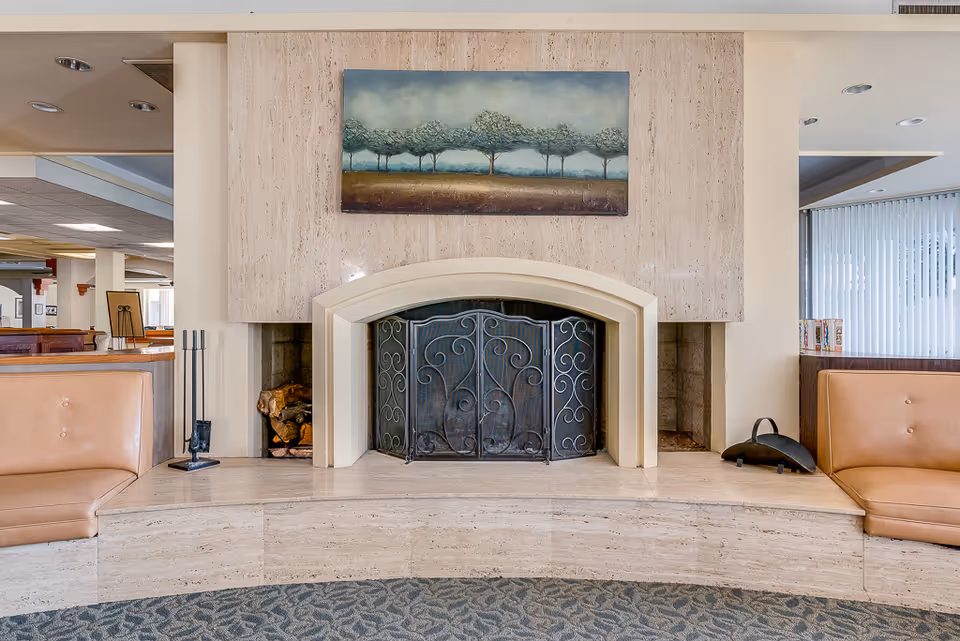 Interior view of a senior living facility lounge area featuring a central fireplace with a decorative metal screen. On either side of the fireplace are built-in seating areas with tan leather cushions. Above the fireplace hangs a painting depicting a row of trees under a cloudy sky. The surrounding area includes carpeted flooring and partial views of the facility's interior with large windows and vertical blinds.