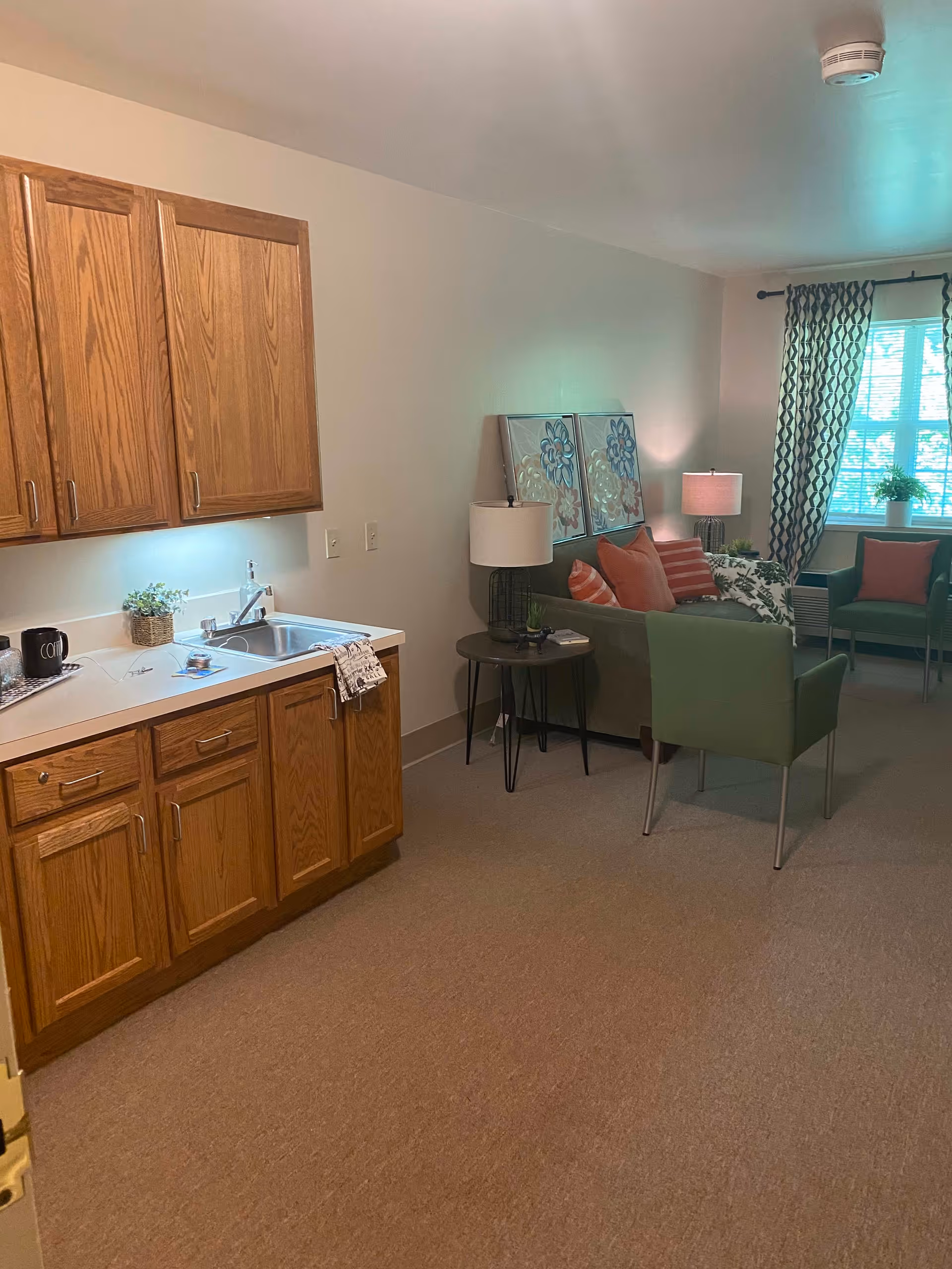 Interior view of a senior living facility room featuring a small kitchenette with wooden cabinets and a sink on the left side. The room also includes a sitting area with a green armchair, a sofa with orange and patterned cushions, two table lamps, framed floral artwork on the wall, and a window with patterned curtains letting in natural light.