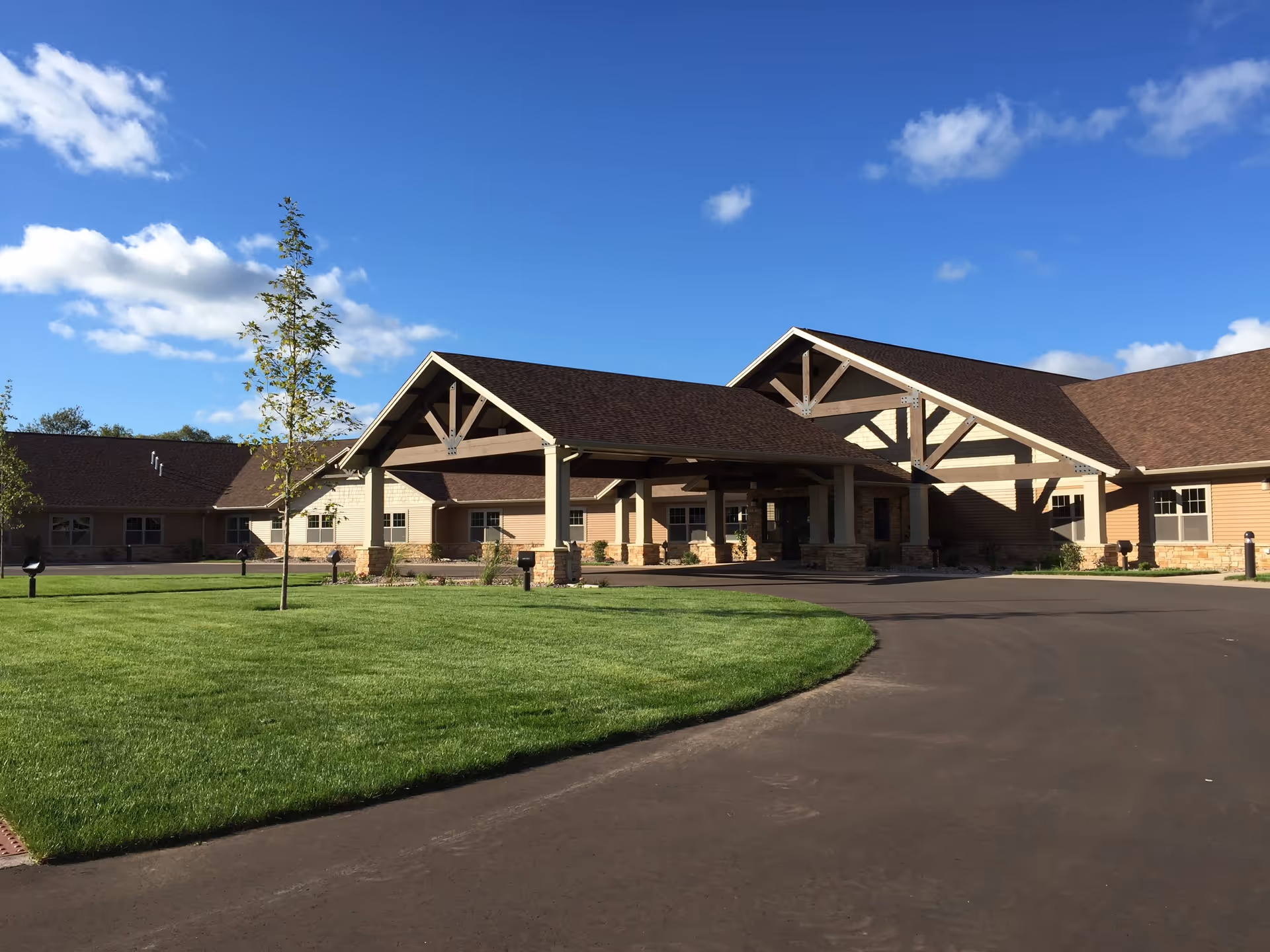 Exterior view of a senior living community building with a large covered entrance, beige siding, stone accents, and a well-maintained green lawn under a blue sky with scattered clouds.