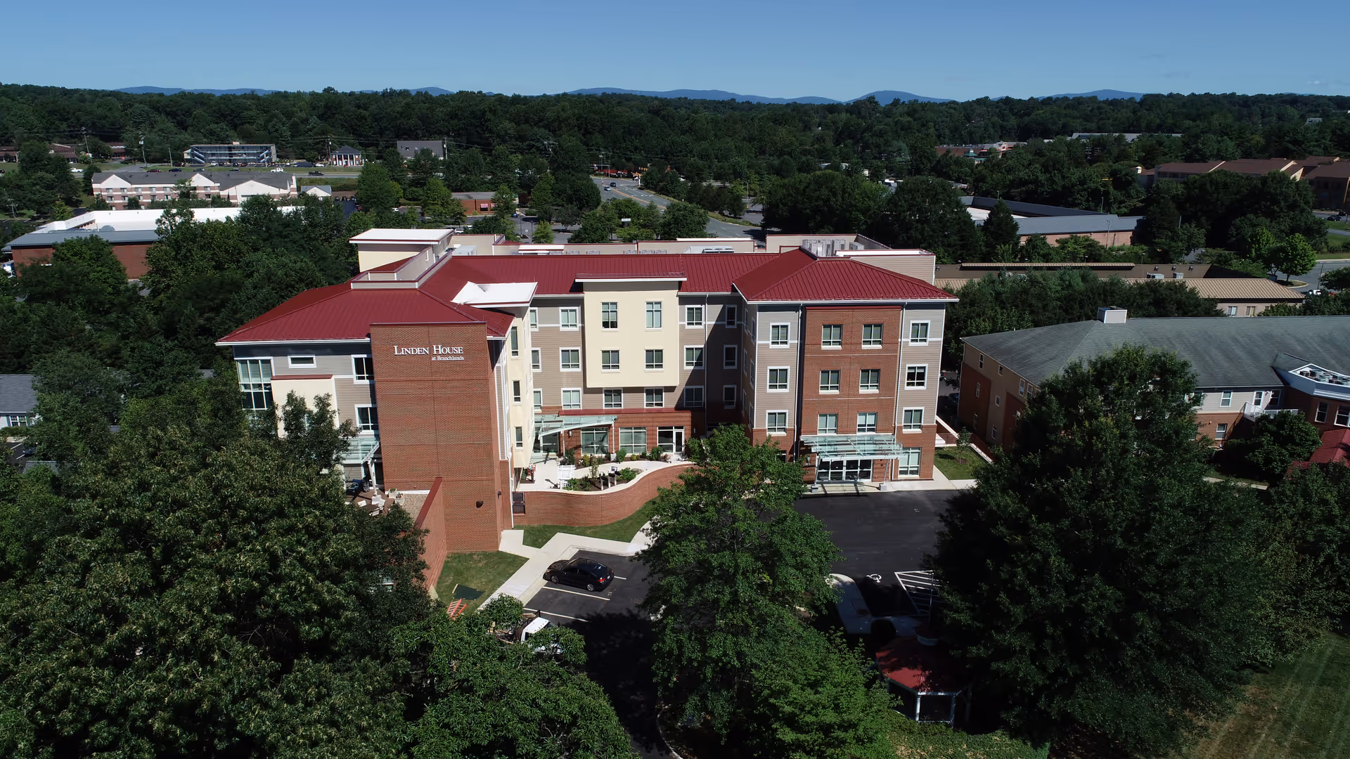 Aerial view of a four-story assisted living building with a red roof, entrance driveway, and surrounding trees.