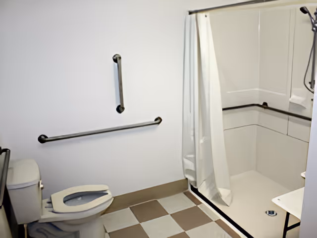 A bathroom with a toilet and a walk-in shower. The shower has a white curtain and grab bars installed on the walls. The floor has a checkered pattern with beige and white tiles.