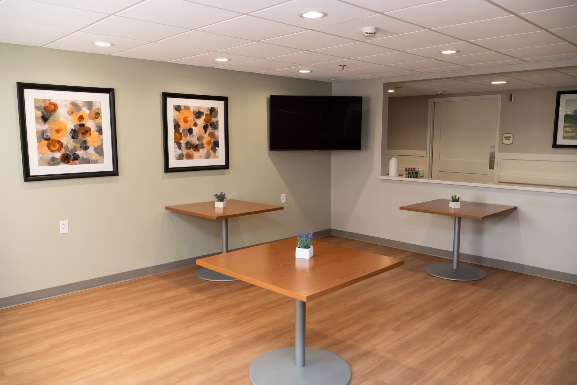 A small common area with three square wooden tables, each with a small potted plant on top. The room has light-colored walls with two framed abstract floral paintings and a flat-screen TV mounted on one wall. The floor is wood laminate, and there is a window opening to another room.