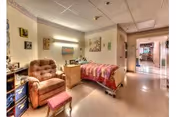 A cozy bedroom in a senior living facility featuring a single bed with a colorful quilt, a cushioned armchair with a matching footstool, a wooden nightstand with decorative items, and artwork on the walls. The room opens into a hallway with additional rooms visible in the distance.