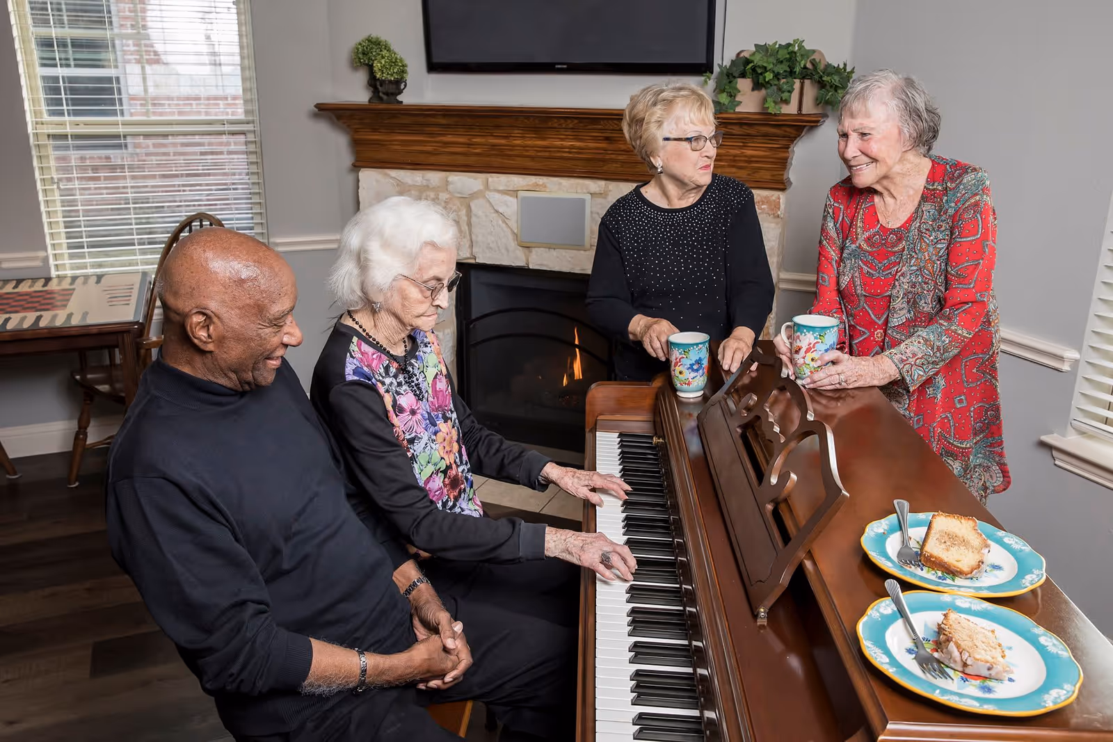 Four elderly people gathered around a piano in a cozy living room. One woman is playing the piano while the others watch and smile. There is a fireplace in the background, two cups on the piano, and two plates with slices of cake on the piano surface.
