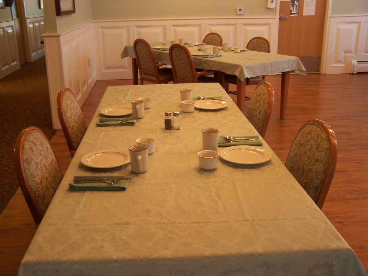 Dining room with two tables covered with light green tablecloths, each set with plates, cups, bowls, silverware, and green napkins. The room has wooden flooring, paneled walls, and upholstered chairs around the tables.