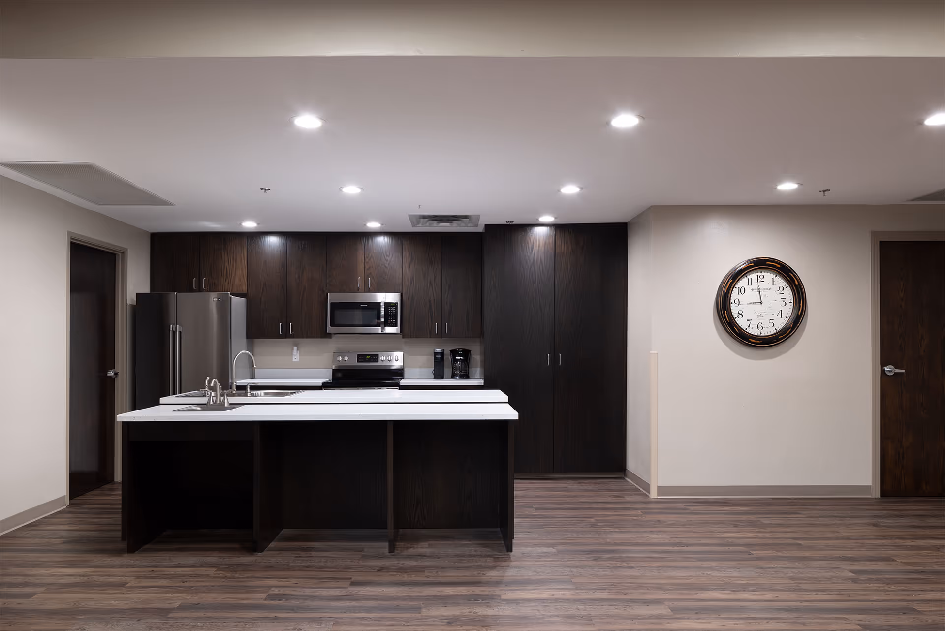 Communal kitchen area with a central island, dark wood cabinetry, stainless-steel appliances, and a wall clock.