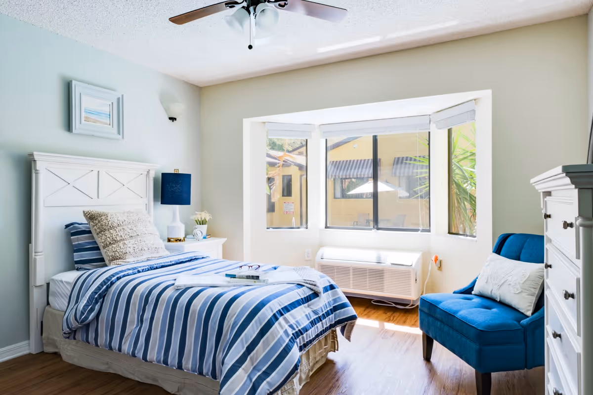 A bright and cozy bedroom with a single bed dressed in blue and white striped bedding, a white headboard, and a beige pillow. Next to the bed is a white nightstand with a blue lamp and a small plant. A blue upholstered chair with a white pillow is positioned near a large bay window that lets in natural light. The room has light-colored walls, wooden flooring, a ceiling fan with lights, and a white dresser on the right side.
