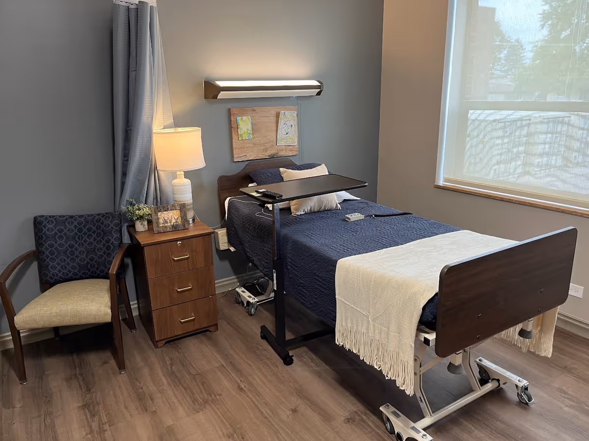 A furnished senior bedroom with a hospital-style bed, overbed table, nightstand with lamp, chair, and a window.