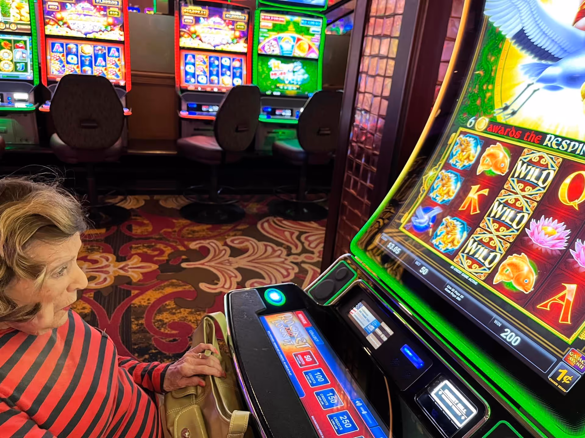An elderly woman in a red and black striped shirt sitting at a slot machine in a casino-like setting with multiple slot machines visible in the background.