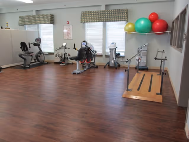 A bright rehabilitation/exercise room with recumbent bikes, therapy equipment, stability balls on a shelf and a wooden gait-training platform on hardwood floors.