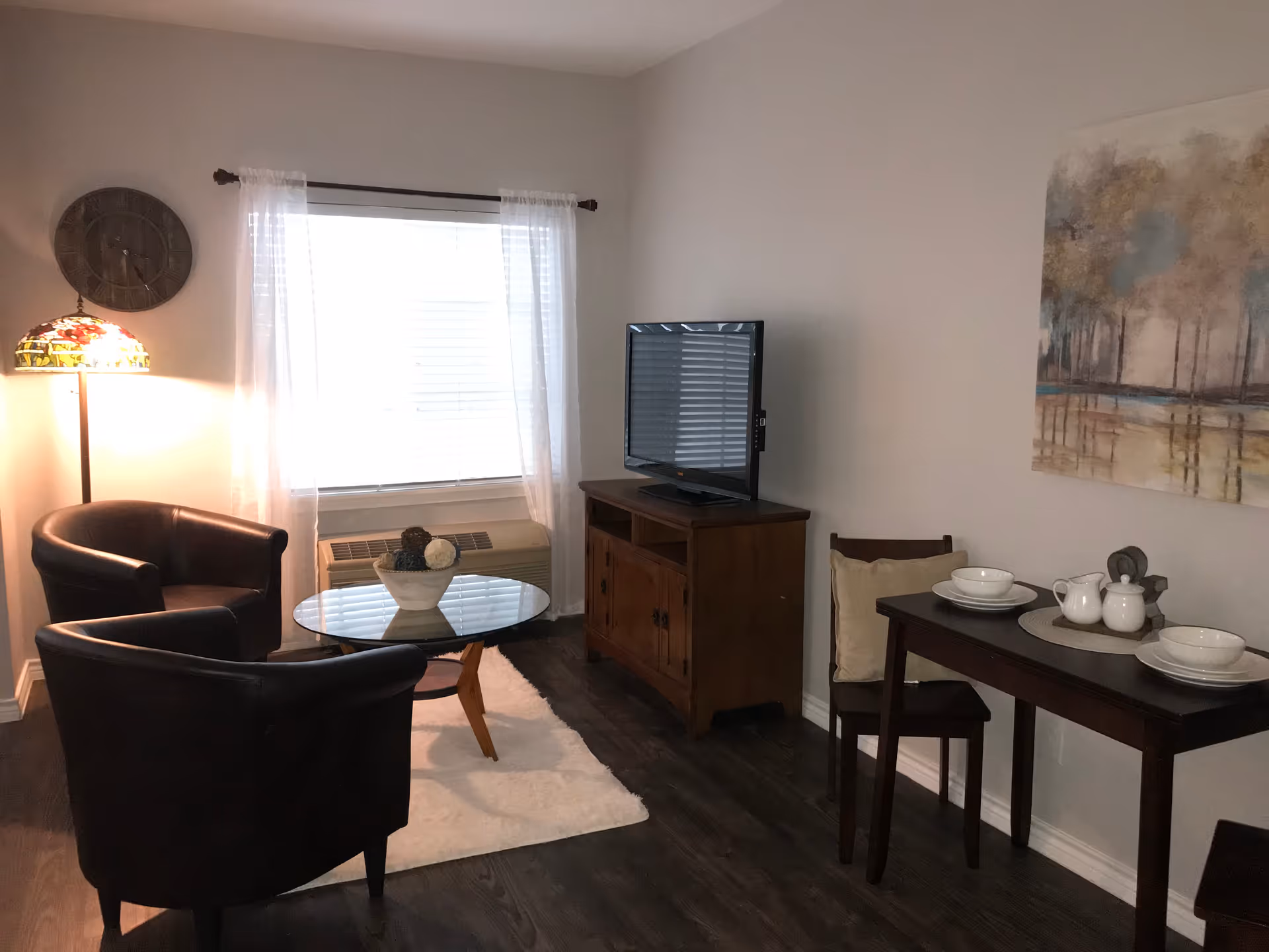A cozy living area with two black leather armchairs around a glass coffee table on a white rug. A wooden TV stand with a flat-screen TV is against the wall. Next to it is a small dining table set with two place settings, a teapot, and cups. A large window with sheer white curtains lets in natural light, and a floor lamp with a stained glass shade stands in the corner. A painting of trees and water hangs on the wall above the dining table.