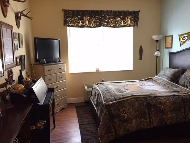 A bedroom with a bed covered in camouflage-patterned bedding, a window with a valance, a tall dresser with a TV on top, a floor lamp, and various decorations on the walls including a Green Bay Packers pennant.